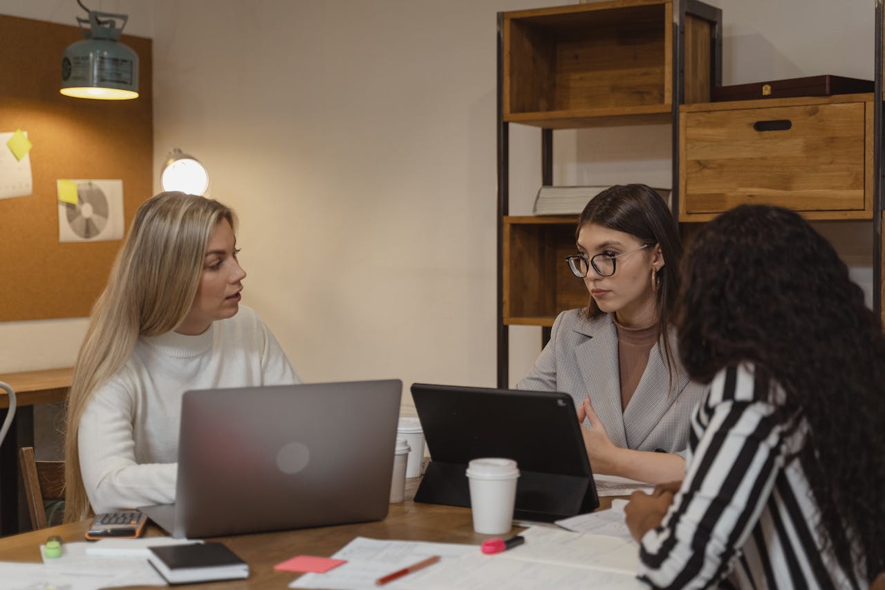 Home Three women engaging in a focused discussion in an office setting with laptops and documents.